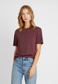 Brombeerfarbenes T-Shirt mit kurzen Ärmeln, aus einem weichen, leichten Stoff gefertigt. Mit rundem Halsausschnitt und lässiger Passform, kombiniert mit hellblauen Jeans.