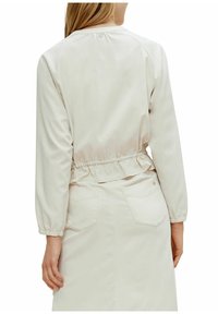 Lys beige jakke med elastisk talje og lange ærmer, der har en knaplukning bagpå og en subtil glans i stoffet.