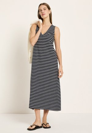 Frau trägt ein ärmelloses, midi-langes Kleid in Marineblau mit weißen horizontalen Streifen, hält eine cremefarbene Jacke über einer Schulter und trägt schwarze Sandalen.