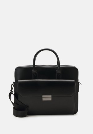 Mallette en cuir noir avec poignées supérieures, poche avant à rabat avec fermoir argenté et bandoulière réglable sur fond blanc.