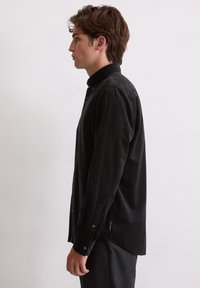 Junger Mann mit braunen Haaren, der ein schwarzes Kordhemd mit Knöpfen und dunkle Hosen trägt, steht im Profil vor einem einfarbigen hellen Hintergrund.