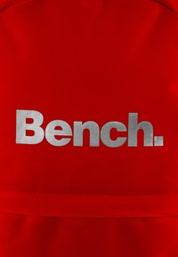 Roter Stoff mit einer glatten Textur, auf dem das silberne Logo "Bench." prominent in der Mitte aufgedruckt ist.