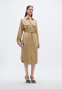 Καφτάνι σε χακί χρώμα με μακριά μανίκια, κουμπιά μπροστά, δύο τσέπες στο στήθος, ζώνη που ταιριάζει και ασύμμετρο τελείωμα. Μοντέλο κρατάει ασημί τσάντα χειρός.