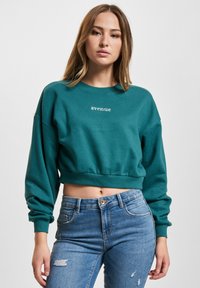 Teal avkortet hettegenser med lange ermer, med "avenue" brodert i hvitt. Kombinert med lyseblå slitne jeans.