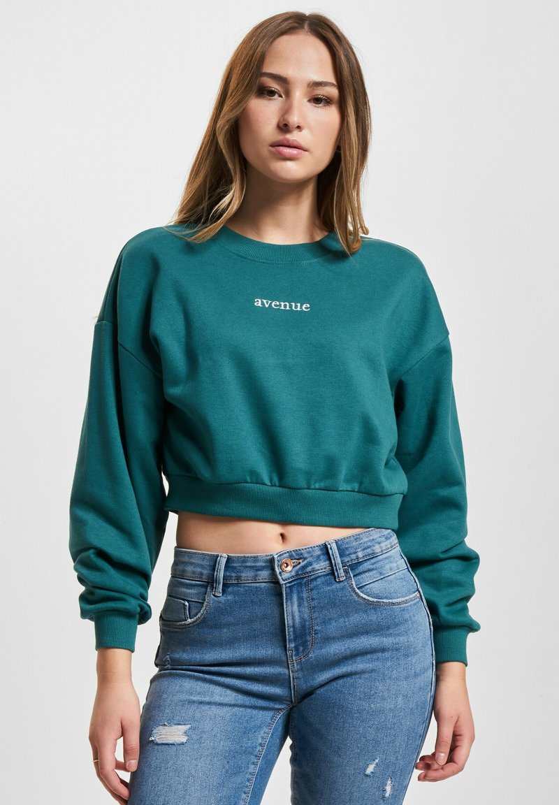Teal avkortet hettegenser med lange ermer, med "avenue" brodert i hvitt. Kombinert med lyseblå slitne jeans.