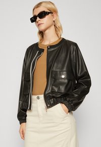 Bomber jacket in ecopelle nera con dettagli bianchi della zip, polsini elasticizzati e due tasche frontali. Top beige aderente e gonna chiara indossati sotto.