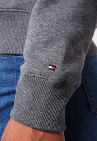 Zbliżenie na szary rękaw swetra dzianego z małym prostokątnym logo w kolorach czerwonym, białym i granatowym nad nadgarstkiem, noszonego z niebieskimi dżinsami.