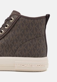 Gros plan de l'arrière et du côté d'une basket montante marron à motifs avec une semelle crème, des œillets pour lacets et « Michael Kors » embossé sur la semelle.