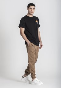 Camiseta negra de cuello redondo con un bolsillo en el pecho, pantalones cargo marrón claro con múltiples bolsillos y zapatillas blancas con acentos en contraste.