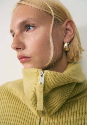 Geel ribbeltrui met een hoge kraag en een zilveren ritsdetail. Model heeft kort, blond haar en gouden creolen.