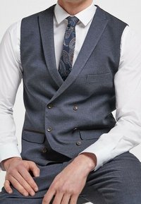 Ciemnoszara dwurzędowa kamizelka z jednym kieszonką, brązowymi guzikami, noszona na białej koszuli, uzupełniona wzorzystym krawatem.