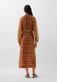 Maxime dress inisi multicolored stripes yenye mshipa wa elastic, mikono mirefu, na undani wa nyuma ulioandaliwa. Imevaajiwa na viatu vya sandali vyenye vidoti vya rangi ya kahawia.