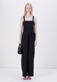 Mono negro con un cuerpo smock ajustado, diseño de pierna ancha y tirantes gruesos. La modelo sostiene un pequeño bolso negro y lleva sandalias sencillas.