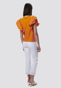 Top corto naranja con volantes y mangas cortas, jeans blancos recortados con detalles de ojales en el dobladillo, y sandalias de tacón alto blancas. Diseño simple, aspecto informal.