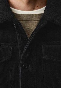 Sort corduroy skjortejakke med en struktureret uldkrave, der har knaplukning og brystlommer, lagd over en beige sweater.