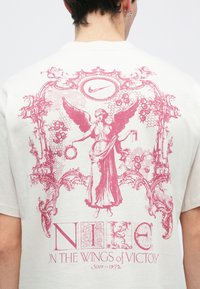 Λευκό μπλουζάκι με μεγάλο ροζ γραφικό ενός φτερωτού προσωπικού με κείμενο "NIKE ON THE WINGS of VICTORY" σε διακοσμητικό, vintage στυλ.