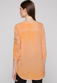 Orange, halbtransparente Bluse mit Dreiviertelärmeln, die dekorativen Text auf dem linken Ärmel und einen abgerundeten Saum aufweist. Weiches Material.