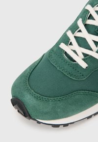 Zapatillas verdes con parte superior de ante y textil, cordones blancos y una suela de goma negra contrastante con tracción texturizada.