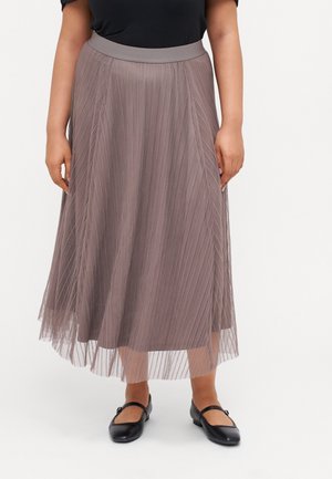 Femme portant une jupe midi plissée taupe avec une taille haute et des chaussures Mary Jane noires, debout sur un fond blanc.
