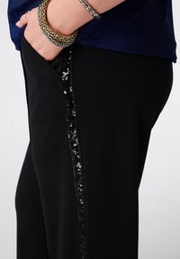Zwarte broek met een zijstreep van zwarte pailletten. Materiaal lijkt zacht en rekbaar. De hand van het model rust in een zak, waarbij accessoires worden getoond.