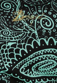 Musta kangas, jossa on turkooseja abstrakteja kukka- ja paisley-kuvioita, ja ylhäällä keskiosassa on kultakirjaillun kursiivitekstin sanotaan.