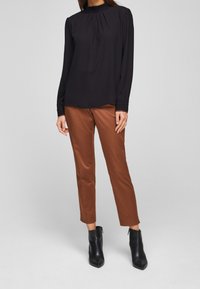 Schwarze Bluse mit langen Ärmeln und gepufftem Ausschnitt, kombiniert mit braunen, eng anliegenden Hosen und schwarzen Stiefeletten mit spitzen Zehen.
