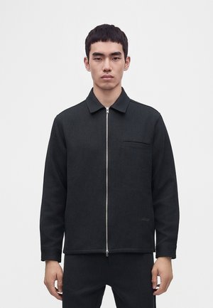 Junger Mann trägt eine schwarze Reißverschlussjacke mit Kragen und Brusttasche, steht vor einem einfarbigen hellen Hintergrund und schaut nach vorne.