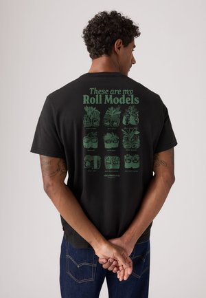 Homme aux cheveux bouclés debout, les mains derrière le dos, portant une chemise noire avec des illustrations de rouleaux de sushi verts et texte "These are my Roll Models."