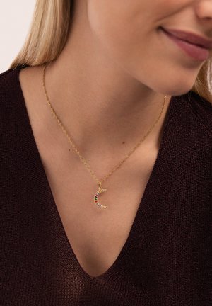 Collar de oro con un colgante de luna creciente adornado con piedras de colores, llevado sobre una camiseta de escote en v. Cadena delicada con un diseño retorcido.