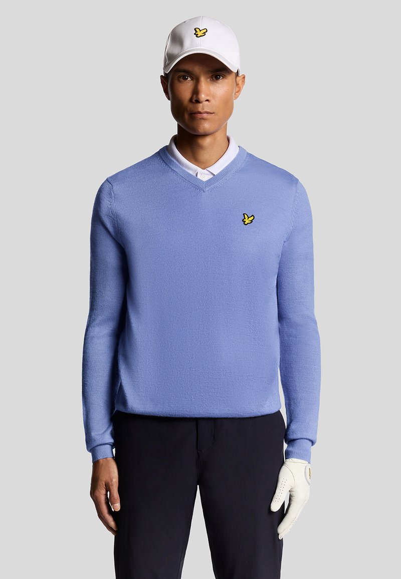 Maglione lavorato a maglia di colore azzurro chiaro con scollatura a V, caratterizzato da un piccolo logo giallo. Indossato con una camicia a colletto bianco e un cappellino bianco, completato da un guanto bianco.