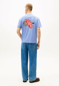 Jasnoniebieska bawełniana koszulka z graficznym wzorem na plecach, przedstawiającym czerwoną metkę i czarną ilustrację. W połączeniu z luźnymi dżinsami.