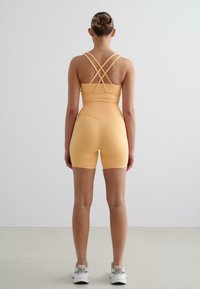Oranje sporttop met kruislings straps en bijpassende shorts. De stof lijkt rekbaar en soepel. Het model draagt witte sneakers.