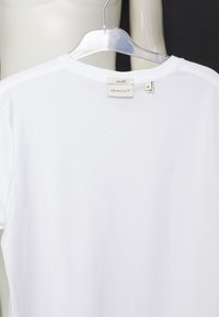 Wit slim fit GANT T-shirt aan een doorzichtige plastic hanger, achteraanzicht met kraag en maat M-label tegen een witte en zwarte achtergrond.