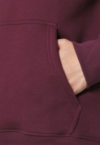 Sudadera burdeos con bolsillo canguro, puños acanalados y textura suave. Incluye un detalle de costura visible a lo largo del bolsillo.