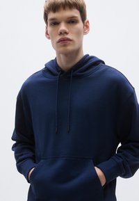 Hanorac albastru marin din material moale, cu glugă ajustabilă prin șnur, buzunar frontal și manșete striate. Logo-ul brandului este afișat discret pe piept.