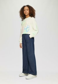 Lichtgekleurde sweatshirt met kleurrijke bloemenpatronen, gecombineerd met donkerblauwe wijde denim broeken en witte sneakers.