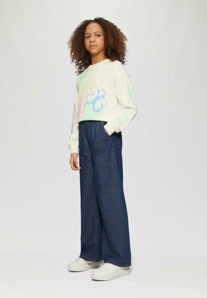 Helle Creme-Sweatshirt mit bunten Blumenmustern, kombiniert mit dunkelblauen, weit geschnittenen Jeanshosen und weißen Sneakers.