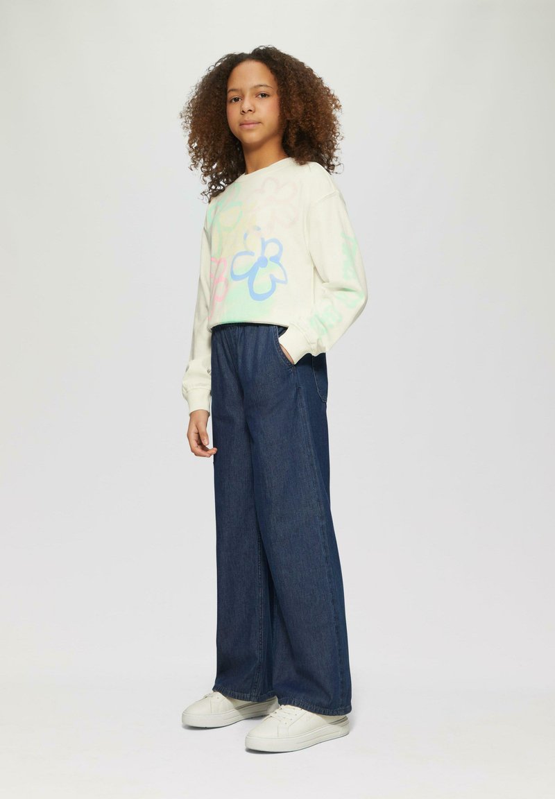 Lichtgekleurde sweatshirt met kleurrijke bloemenpatronen, gecombineerd met donkerblauwe wijde denim broeken en witte sneakers.