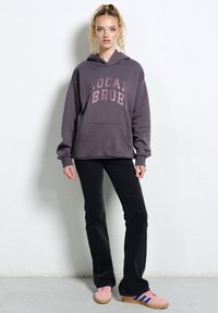 M�łoda kobieta stoi ubrana w ciemną, oversize'ową bluzę z kapturem z napisem "LOCAL ERROR", czarne rozszerzane spodnie oraz różowe sneakersy z niebieskimi paskami.