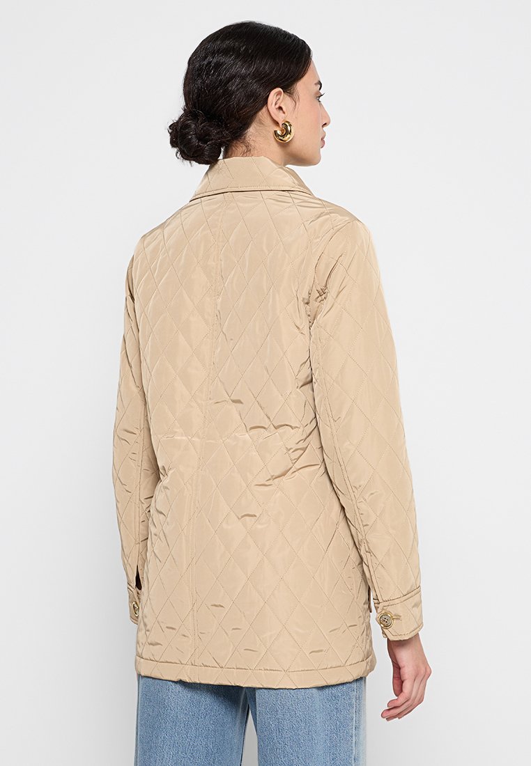 Giacca trapuntata beige con collo a camicia, maniche lunghe e polsini abbottonati. Presenta una superficie testurizzata e un design a taglio dritto.