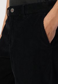 Sorte fløjl bukser med en slim fit, der har lodrette ribber, sidelommer og en metalknaplukning i taljen.