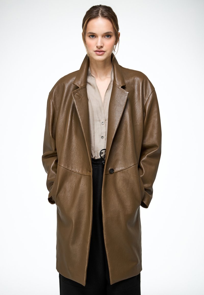 Kobieta w luźnym, brązowym skórzanym płaszczu na beżowej bluzce i czarnych spodniach, stojąca na tle jednolitej białej ściany.