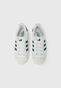 Baltas sporta kurpes ar zaļām svītrām, apvilkts purngala dizains, šņoru aizdare un teksturēta gumijas zole. Zīmola logotips redzams uz mēlītēm.