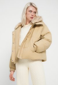 Μπεζ μπουφάν με πούπουλο και κουκούλα με γούνα, διαθέτει πλαϊνές τσέπες, ελαστικά μανίκια και κλείσιμο με φερμουάρ μπροστά, συνδυασμένο με κρεμ πλεκτό πουλόβερ με σχέδιο κορδονιού.