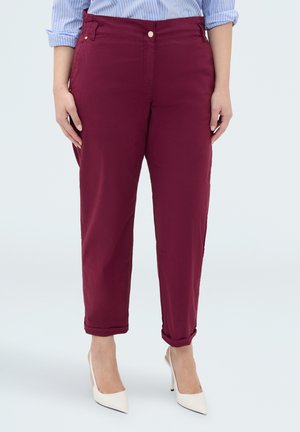 Kvinde iført opkrebede burgunderbukser, lyseblå stribet skjorte og hvide spidse høje hæle, stående foran en ensfarvet baggrund.