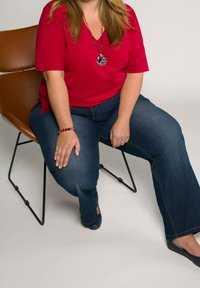 Röd kortärmad topp, blå utsvängda jeans, sittande på en brun stol. Silverfärgad cirkulär hängehalsband och röd pärlhalsband synliga.