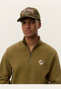 Sweatshirt zippé vert olive avec broderie sur le devant et une casquette assortie olive arborant un texte multicolore. Texture lisse, coupe décontractée.