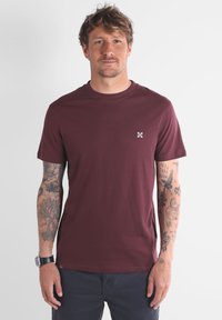 Homme avec des cheveux ébouriffés, portant un t-shirt à manches courtes bordeaux et un pantalon sombre, montrant des tatouages détaillés sur les deux avant-bras.