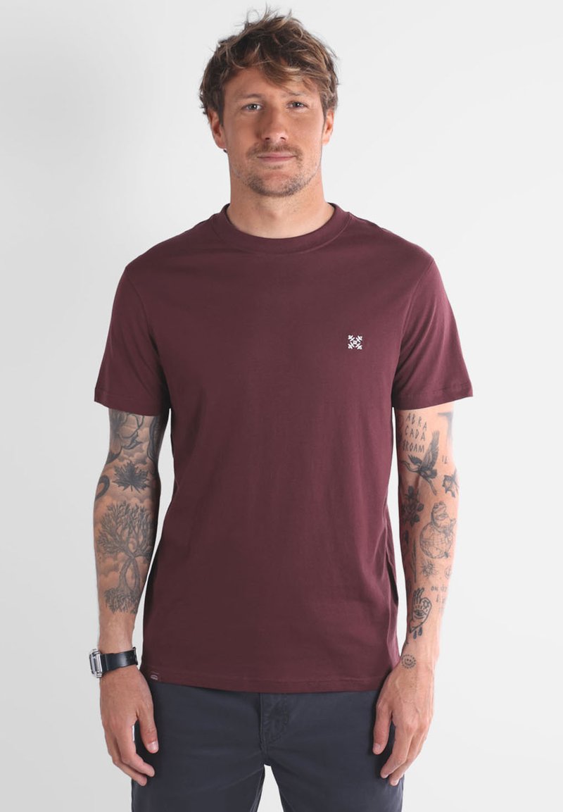 Homme avec des cheveux ébouriffés, portant un t-shirt à manches courtes bordeaux et un pantalon sombre, montrant des tatouages détaillés sur les deux avant-bras.