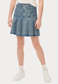 Jupe en denim avec un design plissé, de couleur bleu clair, ourlet effiloché et taille élastique. Portée avec des baskets blanches et des chaussettes côtelées.